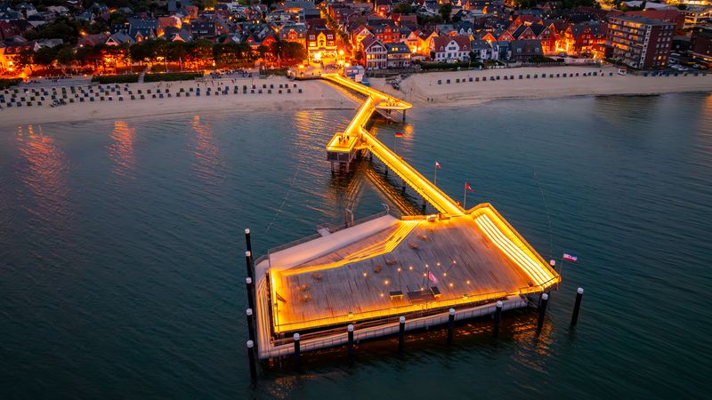 Eine beleuchtete Seebrücke bei Nacht mit Blick auf eine Küstenstadt und den Strand.