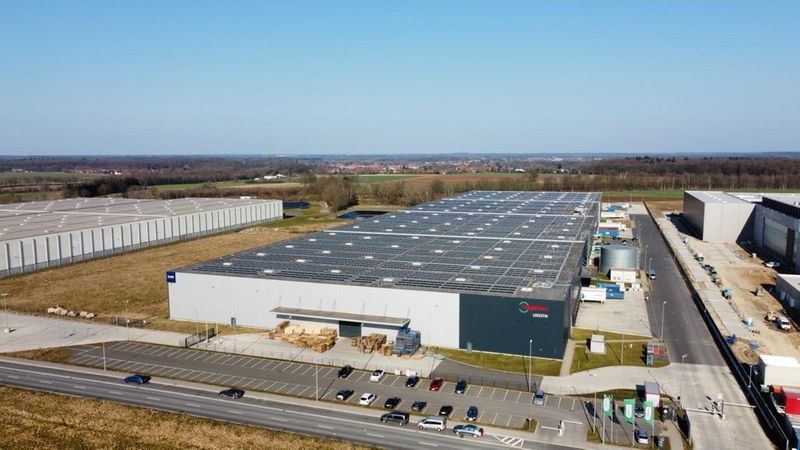 Luftaufnahme eines modernen Logistikzentrums mit Solarpanelen auf dem Dach und umliegenden Parkplätzen.
