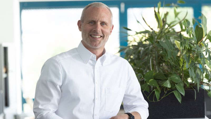 Ein lächelnder Mann in einem weißen Hemd steht in einem Büro neben einer großen Zimmerpflanze.