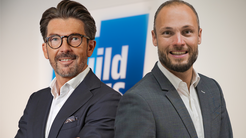 Zwei Geschäftsmänner in Anzügen stehen nebeneinander und lächeln in die Kamera.