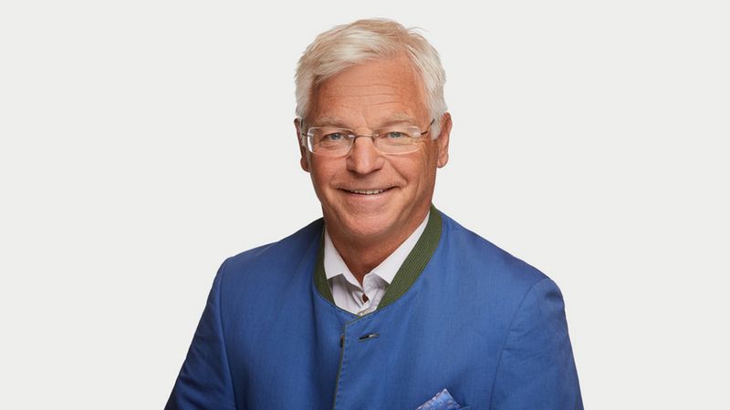 Ein lächelnder älterer Mann in einer traditionellen blauen Jacke vor einem weißen Hintergrund.