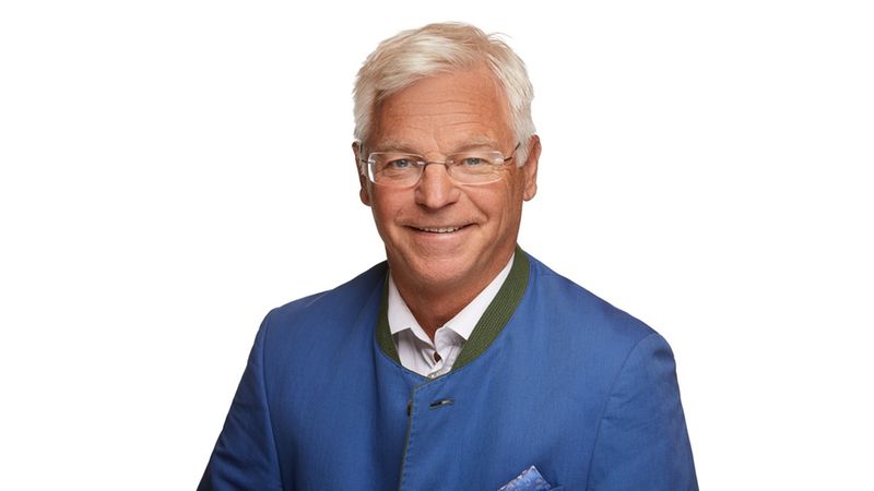 Ein lächelnder älterer Mann in einer traditionellen blauen Jacke vor einem weißen Hintergrund.