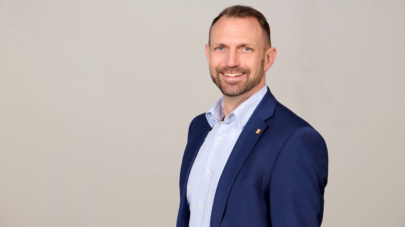 Ein Porträt eines lächelnden Mannes in einem blauen Anzug vor einem neutralen Hintergrund.