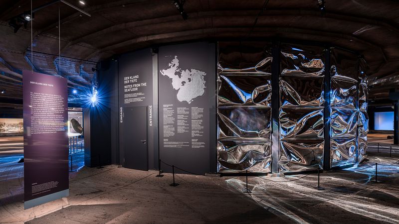 Die Ausstellung "Der Klang der Tiefe" zeigt eine moderne Installation mit reflektierenden Oberflächen und informativen Tafeln.