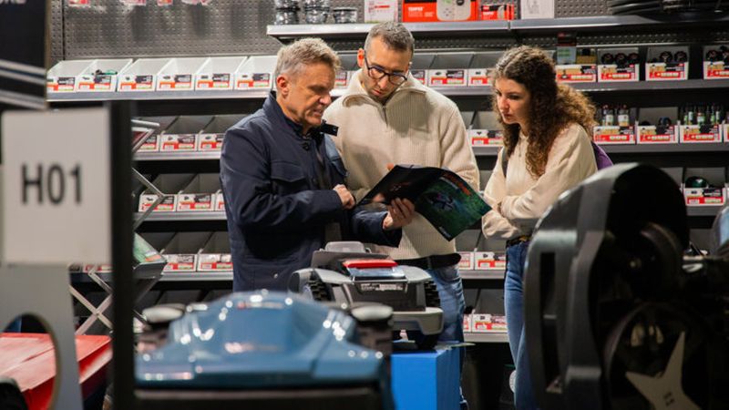 Ein Verkäufer berät zwei Kunden in einem Elektrofachhandel, während sie gemeinsam ein Produktkatalog betrachten.