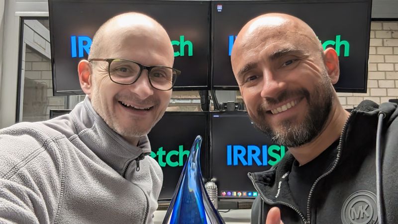 Zwei Personen halten eine blaue Trophäe in einem Büro mit Bildschirmen im Hintergrund.