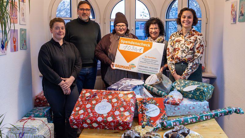 Eine Gruppe von Menschen hält einen großen Spendenscheck in einem Raum mit Geschenken und Gebäck auf einem Tisch.