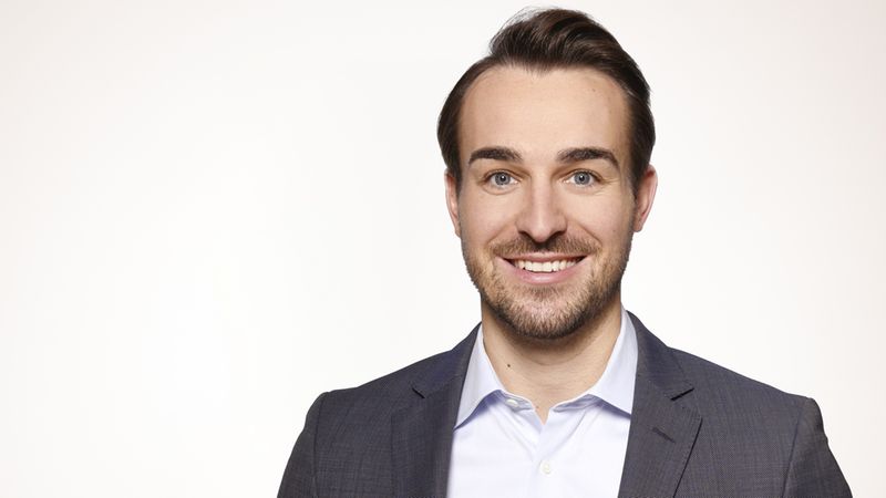 Ein Porträt eines lächelnden Mannes in einem Anzug vor einem neutralen Hintergrund.