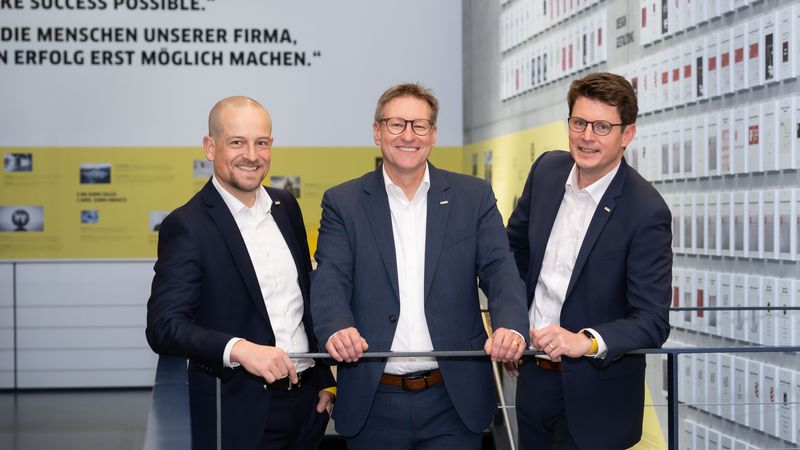 Ein Gruppenfoto von drei Geschäftsleuten in Anzügen, die in einem modernen Bürogebäude stehen.