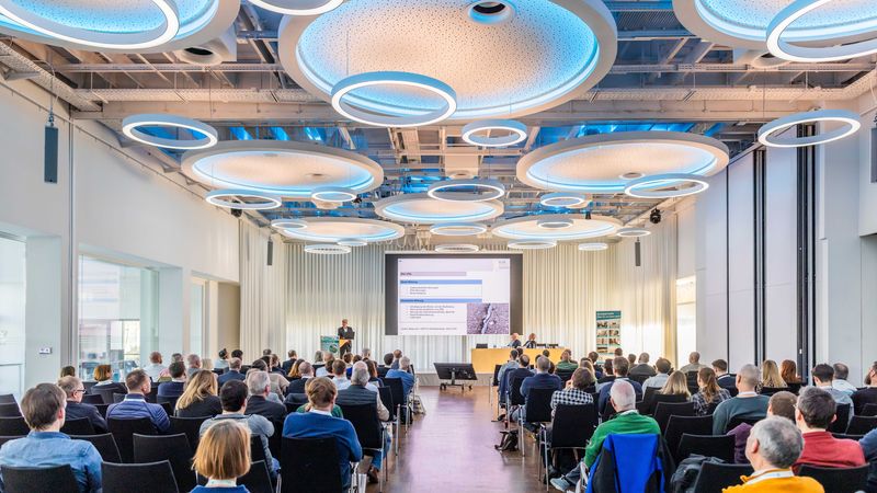 Ein moderner Konferenzraum mit Publikum, das einer Präsentation folgt. Die Decke ist mit futuristischen Leuchten ausgestattet.