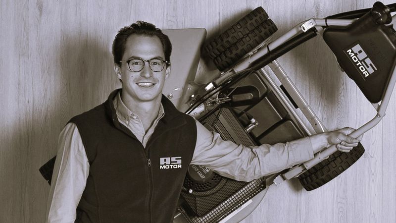 Ein Mann steht neben einem aufgestellten Rasenmäher und lächelt in die Kamera.