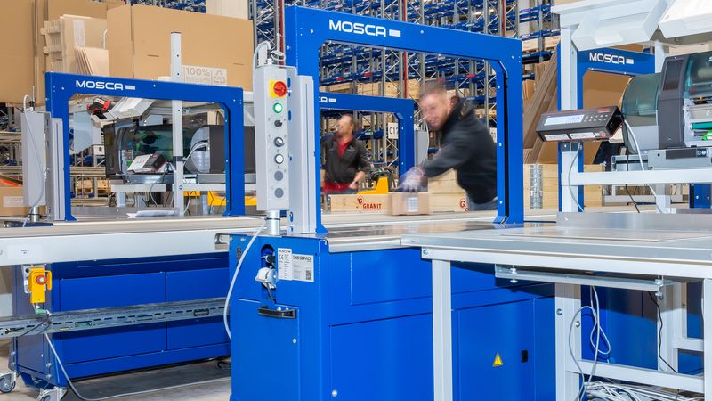 Eine Verpackungsmaschine in einer großen Lagerhalle mit Regalen im Hintergrund.