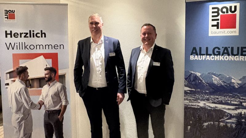 Zwei Männer in Anzügen stehen vor Baumit-Bannern bei einem Baufachkongress.