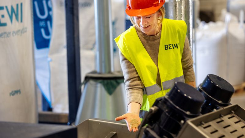 Eine Arbeiterin in einer Fabrik trägt einen roten Schutzhelm und eine gelbe Warnweste, während sie an einer Maschine arbeitet.