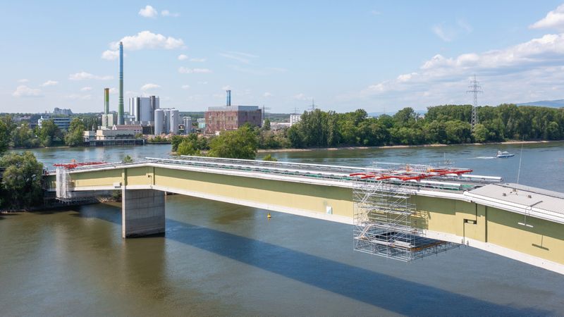 Eine moderne Brücke überquert einen Fluss, im Hintergrund sind Industrieanlagen und grüne Bäume zu sehen.