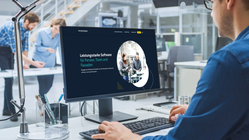 Ein Mann arbeitet an einem Computer in einem modernen Büro, während im Hintergrund Kollegen diskutieren.