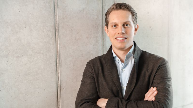 Ein Mann im Anzug steht mit verschränkten Armen vor einer Betonwand.