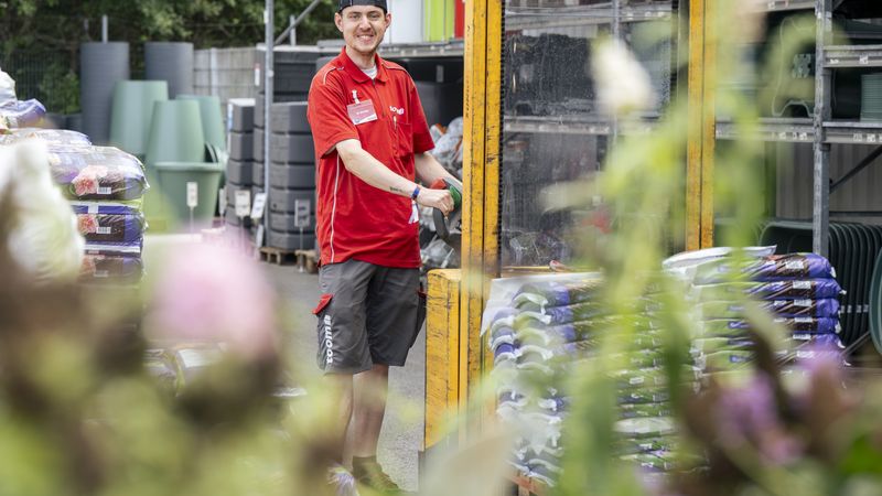 Ein Mitarbeiter in roter Arbeitskleidung steht in einem Gartenmarkt und lächelt in die Kamera. Im Hintergrund sind Pflanzenerde und Gartenzubehör zu sehen.