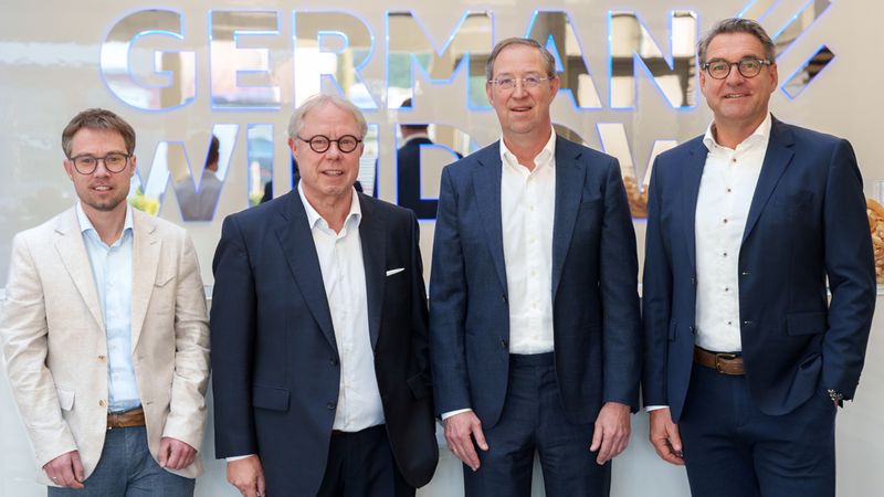 Eine Gruppe von vier Männern in Anzügen steht vor einem Schild mit der Aufschrift "GERMAN WINDOW".