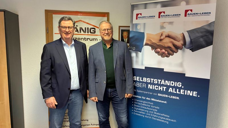 Zwei Männer stehen vor einem Poster mit dem Slogan "Selbstständig, aber nicht alleine" in einem Büro.