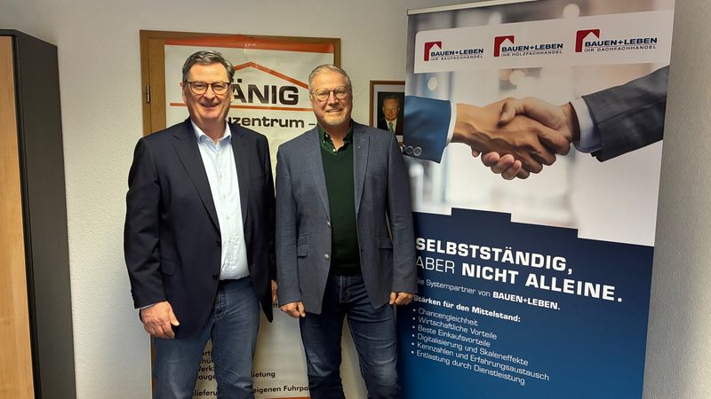 Zwei Männer stehen vor einem Poster mit dem Slogan "Selbstständig, aber nicht alleine" in einem Büro.