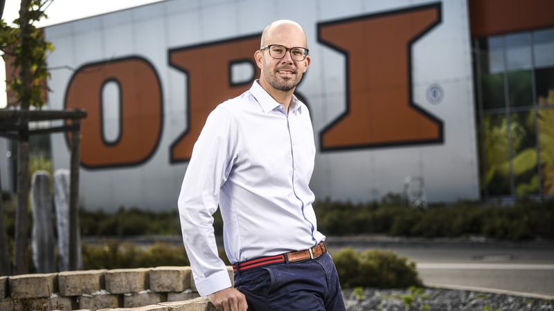 Ein selbstbewusster Geschäftsmann in einem weißen Hemd und einer Hose lehnt entspannt an einer Mauer vor einem modernen Unternehmensgebäude mit großem Logo im Hintergrund.