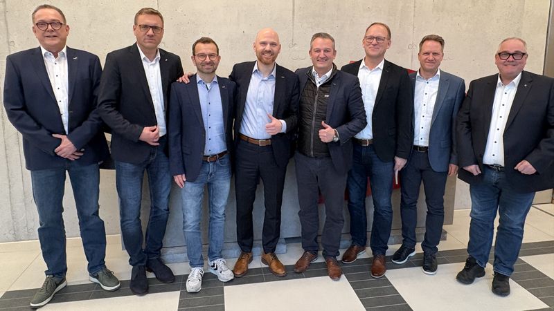 Eine Gruppe von acht Männern in Anzügen steht vor einer Betonwand und lächelt in die Kamera.