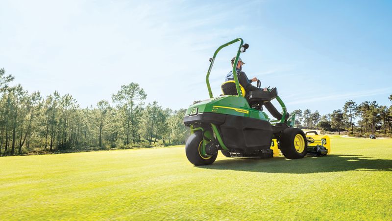 Ein Rasenmäher fährt über einen gepflegten Golfplatz, umgeben von Bäumen und blauem Himmel.