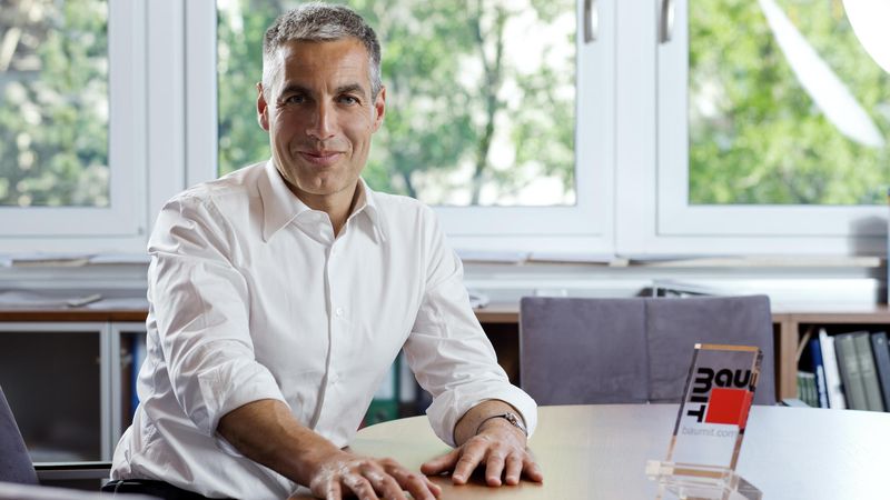 Ein Mann sitzt lächelnd an einem Schreibtisch in einem Büro mit großen Fenstern im Hintergrund.