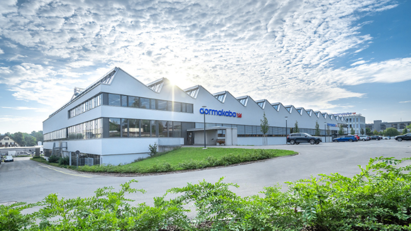 Ein modernes Fabrikgebäude mit Glasdach und großem Parkplatz bei sonnigem Wetter.