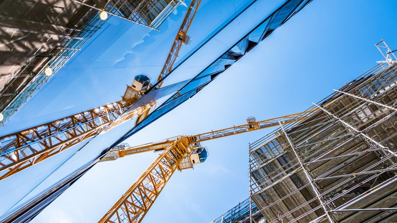 Ein gelber Baukran spiegelt sich in der Glasfassade eines modernen Gebäudes unter einem klaren blauen Himmel.