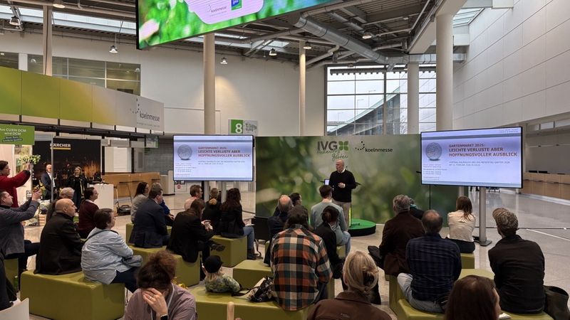 Eine Gruppe von Menschen sitzt in einer Messehalle und hört einer Präsentation zu. Im Hintergrund sind Bildschirme und Banner zu sehen.