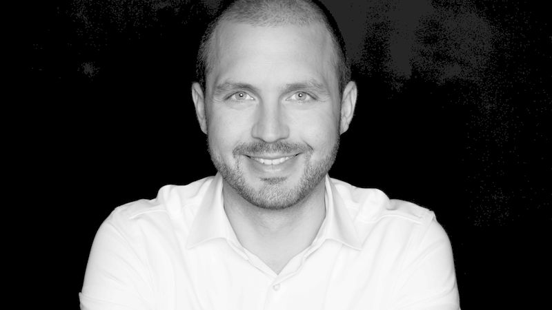 Ein Porträt eines lächelnden Mannes in einem weißen Hemd vor einem schwarzen Hintergrund.