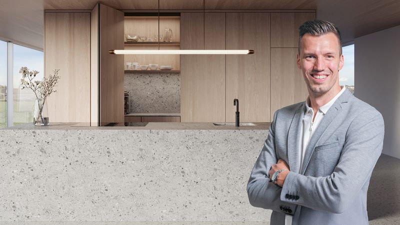 Ein Mann in einem grauen Anzug steht lächelnd vor einer modernen Küche mit Holzverkleidung und einer großen Arbeitsplatte aus Stein.