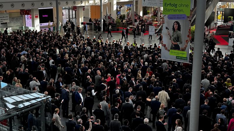 Eine große Menschenmenge in einer modernen Konferenzhalle während einer Veranstaltung. Die Teilnehmer sind geschäftlich gekleidet und stehen unter großen Bildschirmen und Bannern.