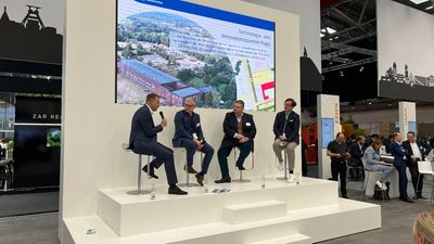 Eine Podiumsdiskussion auf einer Messe, bei der vier Personen auf einer Bühne vor einem großen Bildschirm diskutieren. Der Bildschirm zeigt eine Präsentation mit dem Thema "Technologie- und Innovationsquartier Hugo".