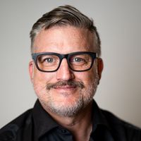 Ein Porträt eines lächelnden Mannes mit Brille und Bart, der ein schwarzes Hemd trägt.