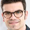 Ein Mann mit Brille lächelt in die Kamera. Der Hintergrund ist unscharf und hell.