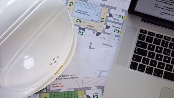 Ein weißer Bauhelm liegt auf einem Tisch neben einem aufgeklappten Laptop, auf dem ein Bauplan ausgebreitet ist, der verschiedene Raumaufteilungen zeigt.