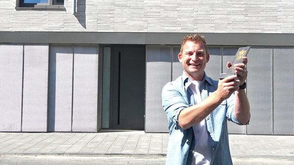 Ein Mann steht lächelnd vor einem modernen Gebäude und hält eine Trophäe in der Hand.