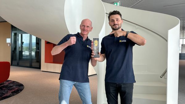 Zwei Personen stehen lächelnd mit einem Pokal in der Hand vor einer modernen Wendeltreppe.