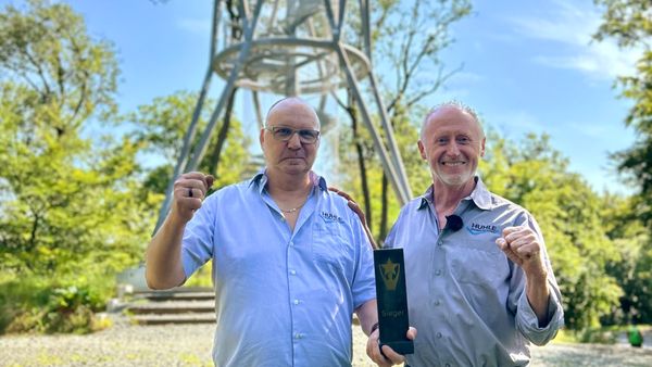 Zwei Männer feiern vor einem großen, modernen Strukturturm in einer grünen Umgebung. Einer der Männer hält eine Trophäe mit der Aufschrift "Sieger".