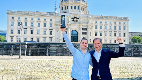 Zwei Personen jubeln vor einem historischen Gebäude, der eine hält eine Auszeichnungstrophäe in die Luft.