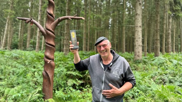 Ein lächelnder Mann im Kapuzenpullover steht in einem dichten Wald und hält eine Auszeichnung. Im Hintergrund ist eine gedrehte Holzskulptur zu sehen.