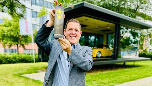 Ein Mann hält stolz einen Pokal in die Höhe, während er auf einer grünen Wiese steht. Im Hintergrund ist ein modernes Gebäude zu sehen.