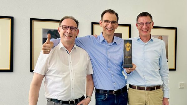 Drei Männer stehen lächelnd nebeneinander, während sie stolz einen Pokal halten.