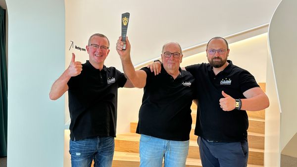 Drei Männer in schwarzen Hemden stehen auf einer Treppe und halten eine Trophäe in die Luft.