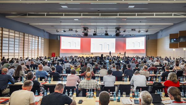 Eine weitwinklige Aufnahme eines modernen Konferenzsaals, in dem zahlreiche Teilnehmer auf Stühlen sitzen und Präsentationen auf einer großen Bühne mit rotem Hintergrund verfolgen.