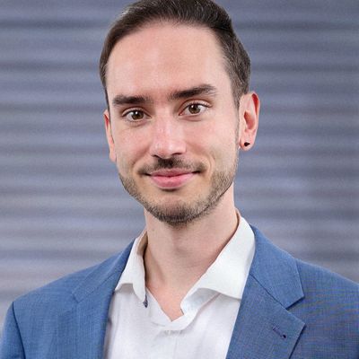 Ein junger Mann in einem blauen Anzug und weißem Hemd lächelt in die Kamera.