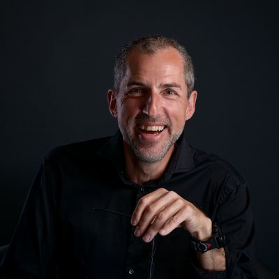 Ein Porträt eines lachenden Mannes in schwarzem Hemd vor einem dunklen Hintergrund.